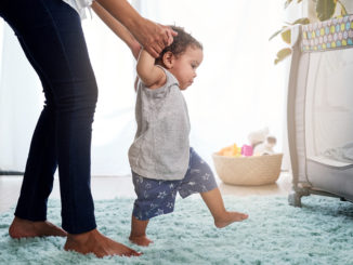 Babys erste Schritte, die Mutters Hände halten, niedliches instabiles Gehen im Kinderzimmer mit Kinderbett